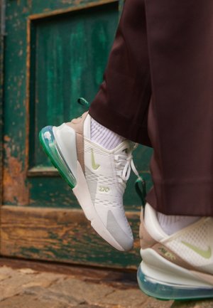 Personne portant des baskets Nike Air Max 270 blanches et beiges avec des touches vertes, debout sur une surface en bois texturée près d'une porte verte usée.