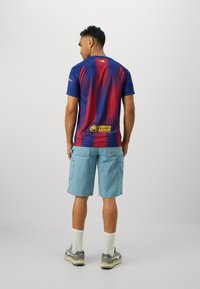 Tricoul de fotbal Nike Barcelona, cu un gradient albastru și roșu, mâneci scurte și logo-ul UNHCR proeminent pe spatele inferior. Purtați-l cu pantaloni scurți din denim.