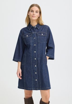 Kvinde iført en mørkeblå denimkjole til knæet med knapper foran, brystlommer og trekvartærmer, stående med den ene hånd i lommen.
