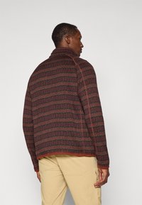 Textured fleece jacket with a high collar, featuring a zigzag pattern in red, green, and black, finished with contrasting trim.