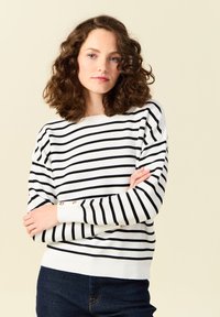 Suéter blanco con rayas horizontales negras. Presenta un cuello barco, puños acanalados y una textura de punto. Estilizado con jeans oscuros.