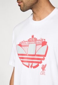 Valge T-särk, millel on suur punane graafiline kujundus tekstiga "Sa ei kõnni kunagi üksi" ja reljeefne vappilaadne logo.