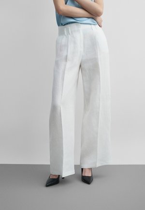 Person trägt weiße Hosen mit weitem Bein und schwarze spitze Absatzschuhe, steht mit verschränkten Armen vor einem schlichten Hintergrund.