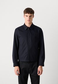 Casaco azul-marinho com fecho éclair e colarinho, feito de tecido suave. Apresenta mangas compridas e um design de corte reto, combinado com calças pretas.