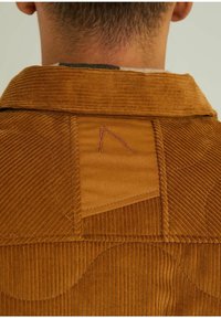 Vue de dos d'une personne portant une veste en velours côtelé marron avec des motifs géométriques cousus et un petit motif brodé au niveau du col.