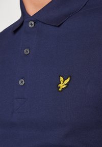 Polo shirt marine en tissu texturé avec une patte de boutonnage à deux boutons et un logo d'oiseau brodé jaune sur la poitrine.