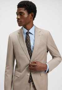 Abito beige chiaro con una texture liscia, caratterizzato da un singolo bottone e rever a punta. Abbinato a una camicia azzurro chiaro e una cravatta marrone a fantasia.