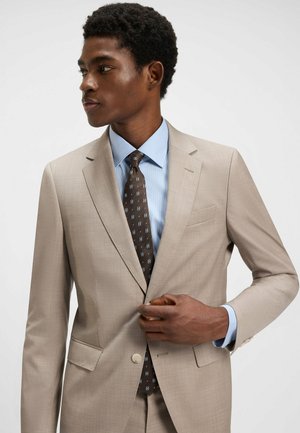 Helle beige drakt med en jevn tekstur, utstyrt med én knapp og hakkede slag. Kombinert med en lyseblå skjorte og et mønstret brunt slips.