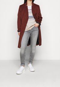 Abrigo largo burdeos de textura suave, combinado con un suéter ligero con patrones geométricos y jeans skinny grises. Zapatillas blancas completan el look.