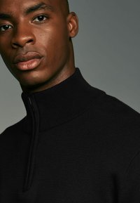 Schwarzer Zip-Up-Pullover mit hohem Rippenkragen, aus strukturiertem Strickstoff gefertigt. Nahaufnahme, die sich auf den Kragen und den Brustbereich konzentriert.