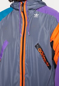 Grå jacka med huva, försedda med orange och lila detaljer, en frontdragkedja och en logopatch. Tillverkad av glatt, lätt material.