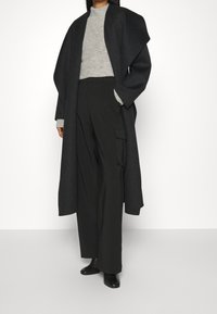Langer, dunkelgrauer Wollmantel, kombiniert mit einem hellgrauen Strickpullover und weit geschnittenen schwarzen Hosen. Hat einen lässigen Schnitt und dezente Texturen.