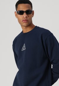 Sudadera azul marino con cuello redondo y gráfico triangular blanco. Modelo usando gafas de sol negras, mostrando un ajuste casual y una textura suave.