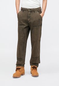 Dark green, relaxed-fit pants with contrast stitching, side pockets, and reinforced knee panels, paired with tan boots.