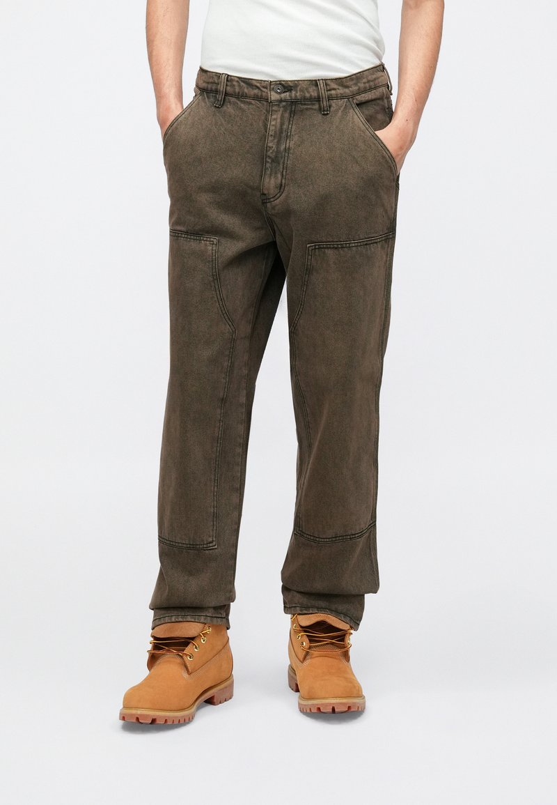 Dark green, relaxed-fit pants with contrast stitching, side pockets, and reinforced knee panels, paired with tan boots.