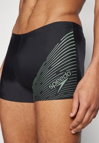 Črne kopalne hlače iz gladkega materiala z zelenim linearjem in logotipom Speedo na stegnu. Oblikovane za zmogljivost in udobje.