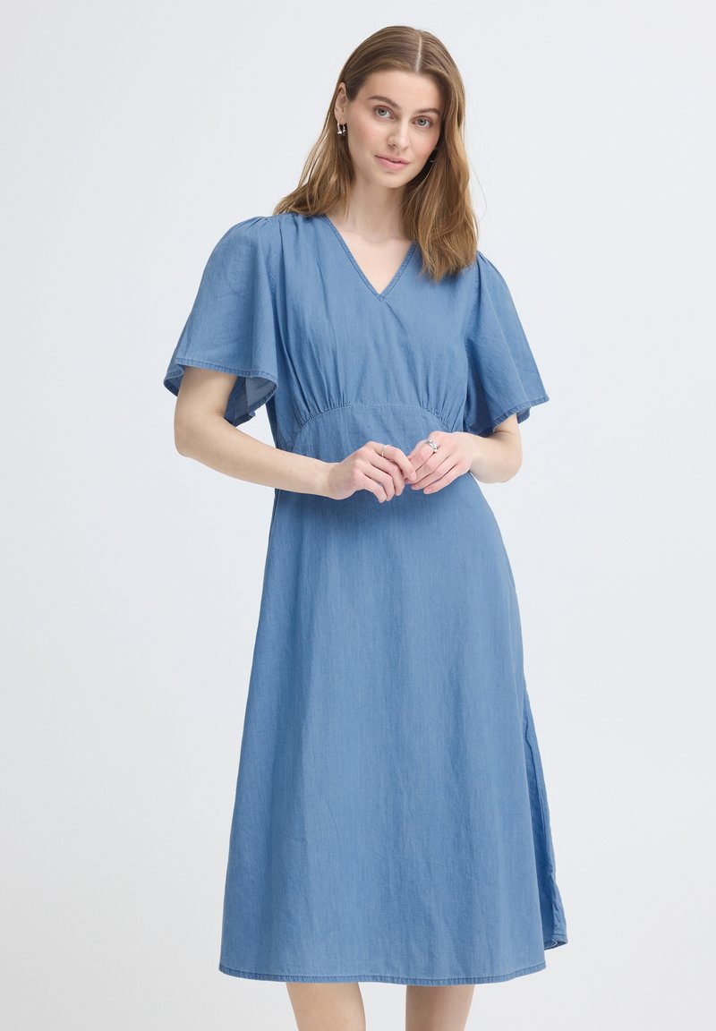 Femme aux cheveux châtain clair portant une robe midi bleue à manches volantées, col en V et fente latérale, posant devant un fond clair uni.