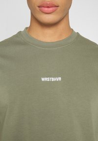 Olivově zelené bavlněné tričko s kulatým výstřihem, s bílým vyšívaným nápisem "WRSTBHVR" na hrudi. Hladká, přiléhavá textura.