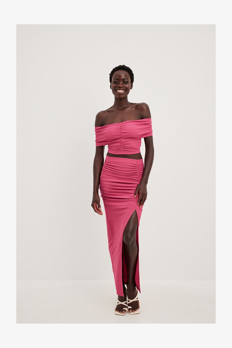 Pink, two-piece outfit featuring an off-the-shoulder top with ruching and a knee-length skirt with a high slit and gathered detailing.