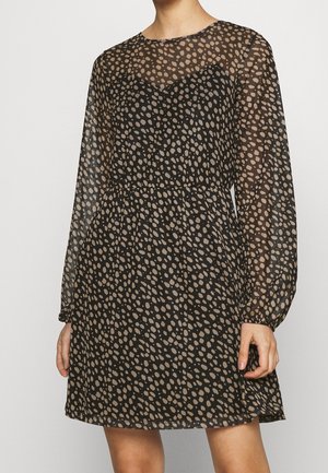 Kvinde iført en sort gennemsigtig kjole med lange ærmer med beige uregelmæssigt prikkemønster og tætsiddende talje.