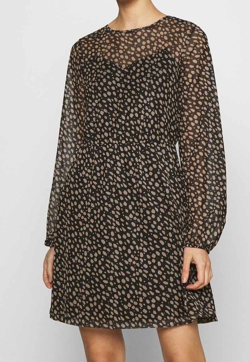 Femme portant une robe noire transparente à manches longues avec un motif de points beige irrégulier et une taille ajustée.