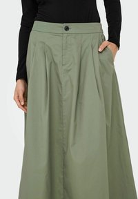 Una gonna plissettata a vita alta in tessuto di cotone verde oliva, dotata di tasche laterali e chiusura con un singolo bottone sul davanti.