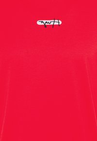 Czerwona koszulka z miękkiego, gładkiego materiału, z czarnym haftowanym logo na białym owalnym tle. Prosty okrągły dekolt i krótkie rękawy.