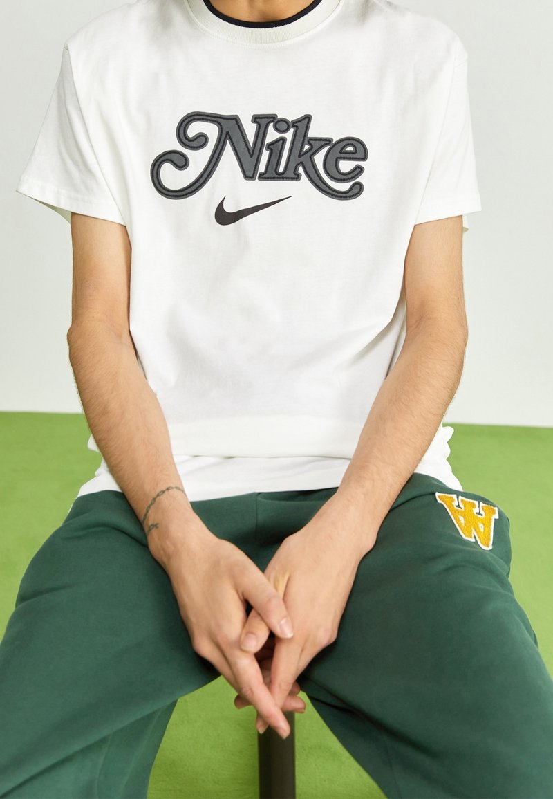 Vit bomullst-shirt med en stor, stiliserad svart Nike-logotyp. Kombinerad med gröna sweatpants som har en gul broderad "W"-detalj.