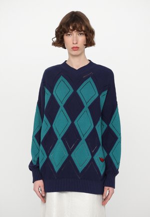 Pull à col en v bleu et turquoise avec un motif en losanges, présentant des poignets et un ourlet côtelés, ainsi qu'un petit détail de logo rouge sur le côté.