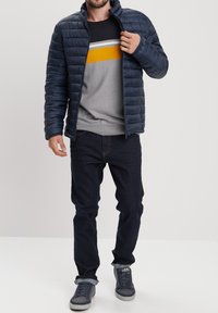 Giubbotto imbottito navy con motivo a quadri orizzontali, indossato sopra un maglione grigio con strisce gialle e grigie, abbinato a jeans scuri e sneakers.
