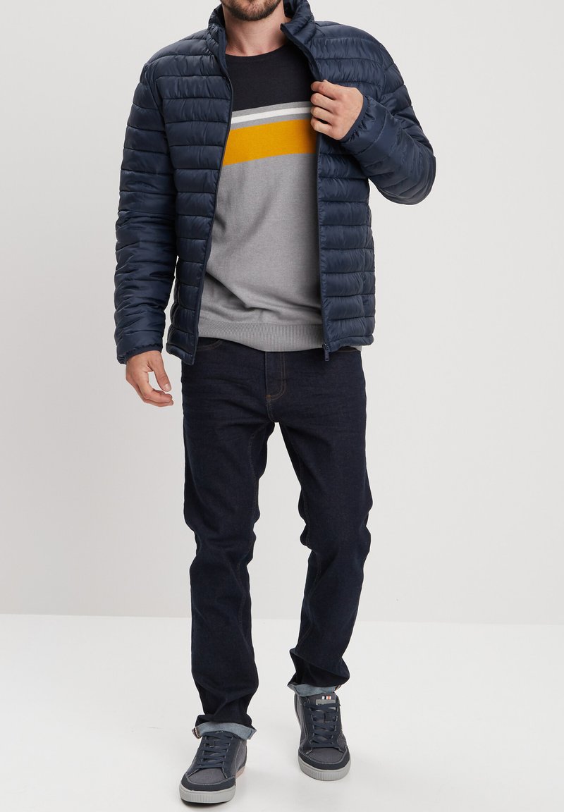 Giubbotto imbottito navy con motivo a quadri orizzontali, indossato sopra un maglione grigio con strisce gialle e grigie, abbinato a jeans scuri e sneakers.