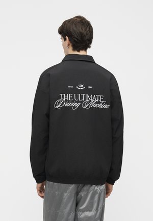 Isik kannab musta jopet, millel on seljal kiri „THE ULTIMATE Driving Machine“, koos läikivate hallide pükstega, seljaga kaamera poole.