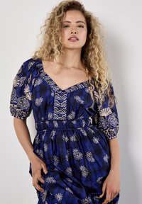 Frau mit lockigem blonden Haar, die ein blaues gemustertes Kleid mit Puffärmeln trägt und vor einer schlichten weißen Wand steht.