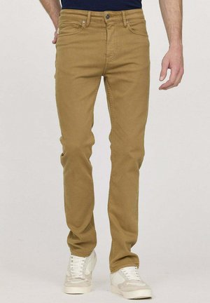 Jean en coton beige avec une coupe ajustée, cinq poches et des accessoires en métal. Porté avec des baskets blanches, affichant une texture lisse et un design épuré.