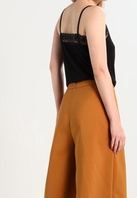 Donna che indossa un top in pizzo nero con spalline e pantaloni larghi a vita alta color senape, in piedi di lato su uno sfondo bianco.