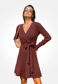Vínová žebrovaná pletená zavinovací šaty s dlouhými rukávy a zavazovacím páskem. Má V-krk a rozšířený lem. Na sobě má neutrální doplňky.