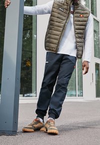Padded olivová vesta přes bílou dlouhou košili, kombinovaná s tmavě modrými tepláky a hnědými botami s oranžovými šňůrkami, stojící venku.