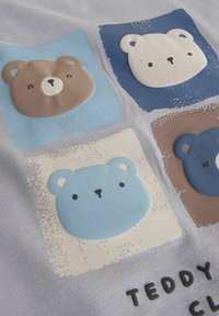 Quatre visages d'ours en peluche dans des carrés de couleur marron, blanc, bleu clair et bleu foncé sur un tissu, avec le mot "TEDDY" partiellement visible en dessous.