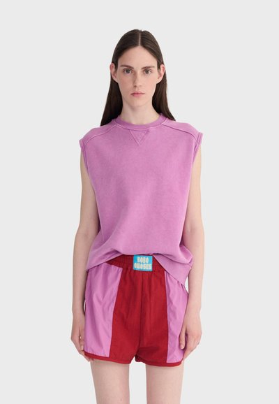 Jeune femme portant un sweat-shirt sans manches violet et un short rouge avec des panneaux latéraux roses et une étiquette bleue, debout devant un fond uni.
