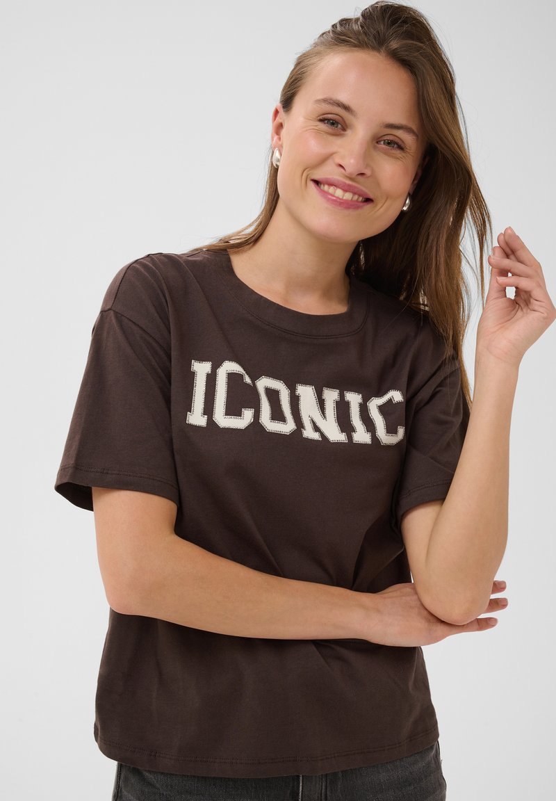 Femme souriante aux cheveux châtain clair porte un T-shirt marron foncé avec le texte « ICONIC », posant avec un bras croisé et l'autre main levée près de sa tête.
