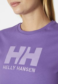 Fialové bavlněné tričko s kulatým výstřihem, velkým bílým logem "HH" a vyšitým textem "HELly HANSEN" níže. Hladká textura.