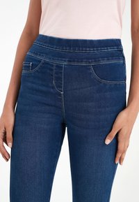 Tmavě modré džíny z denimu s hladkou texturou, s vysokým pasem, předními kapsami a kontrastním šitím. Detailní záběr zaměřený na pas a ruce.