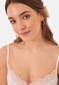 Camisole en satin blanc avec une bordure en dentelle au niveau du col, porté avec des boucles d'oreilles en or. Le modèle a les cheveux bruns et fait légèrement face sur le côté.