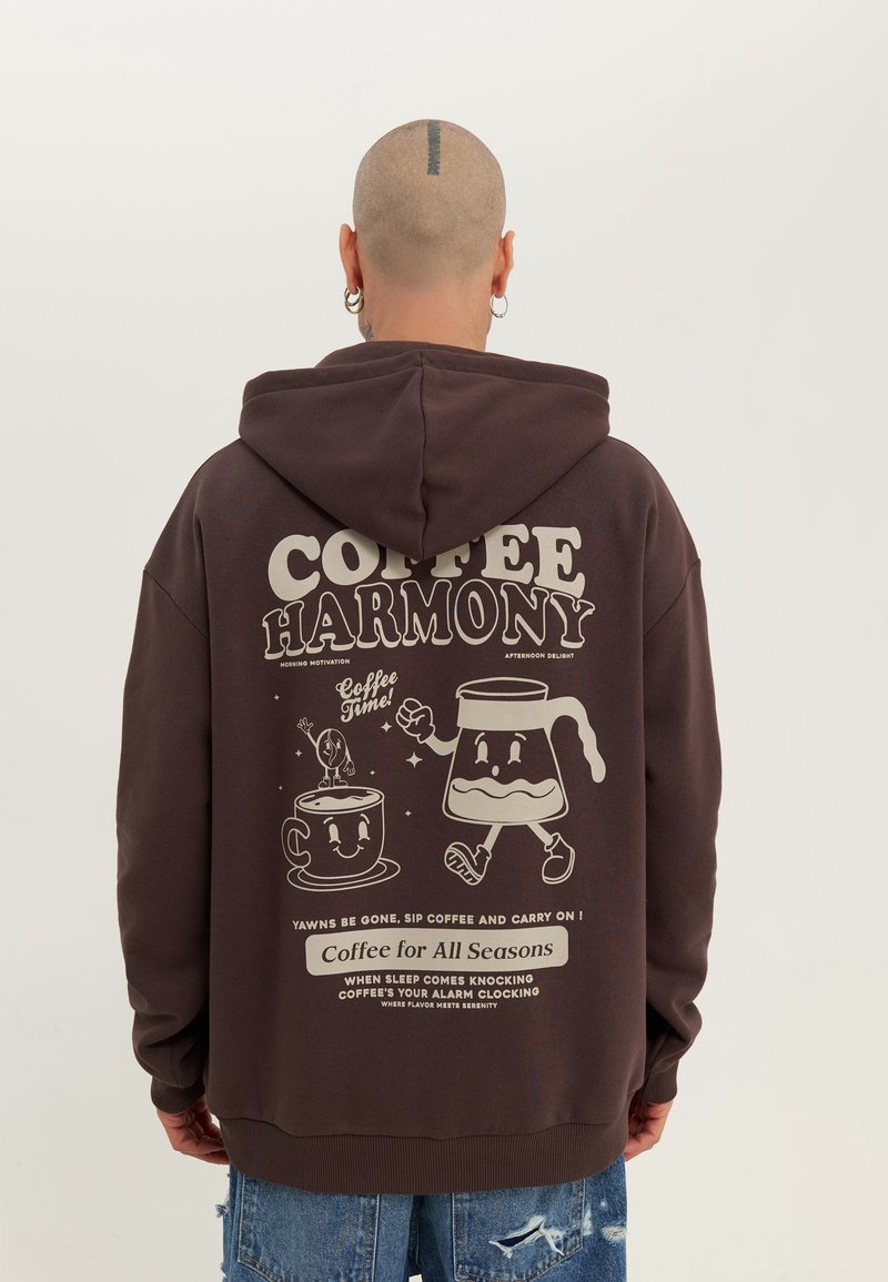 Personne à la tête rasée et aux boucles d'oreilles portant un sweat à capuche sombre avec une tasse de café et une cafetière en dessin animé, ainsi qu'un texte sur le thème du café dans le dos.