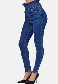 Vaqueros de talle alto en azul con botones en la parte delantera, cinco bolsillos, corte ajustado y un ligero estiramiento. Combinados con tacones negros de punta.
