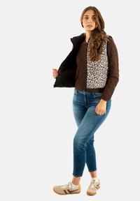 Gilet marrone trapuntato con pannello a stampa animale, indossato sopra una maglietta a maniche lunghe, abbinato a jeans blu e sneaker casual.