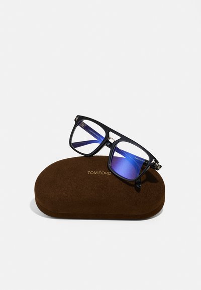 Óculos de sol Tom Ford pretos com lentes azuis repousando sobre uma caixa marrom com textura de camurça, apresentando letras em dourado e um formato arredondado.