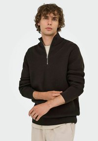 Mörkbrun zipneck-tröja med ribbat mönster och långa ärmar, lagd över en ljusfärgad skjorta. Modell med lockigt hår poserar avslappnat.