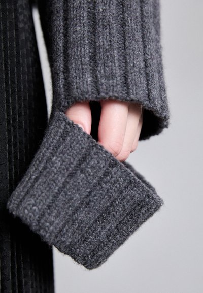 Manga de suéter de malha canelada cinza, com uma mão parcialmente visível segurando o tecido, destacando a textura e um design largo na abertura.
