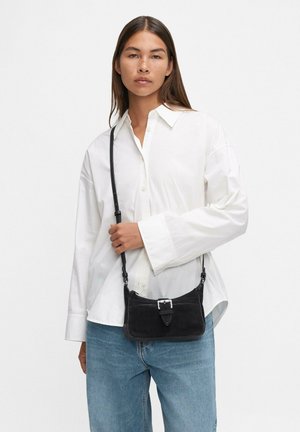 Borsa a tracolla in suede nero con tracolla regolabile e dettagli in bucato argento. Indossata sopra una camicia bianca oversize e jeans azzurri chiari.